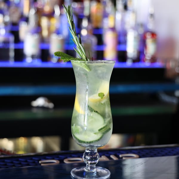 Refreshing cocktail with cucumber, lemon, mint, and rosemary on a bar with blurred bottles.