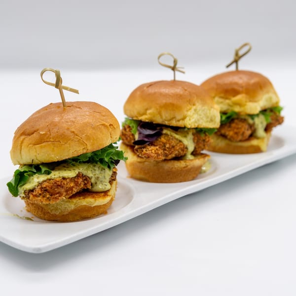 Three crispy chicken sliders with lettuce and green sauce on a white rectangular plate.